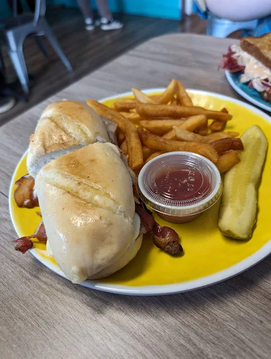 Sandwich on a yellow plate served with fries, a pickle spear and dipping sauce